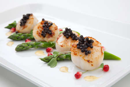 Close up of delicious appetizer scallops with black caviar. Garnished with asparagus, pomegranate seeds, microgreens, and sauce. Elite food.の写真素材