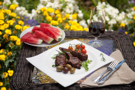 Grilled lamb chops served with cherry tomatoes and olives. Watermelon and wine. Outside dinner in a garden.の写真素材