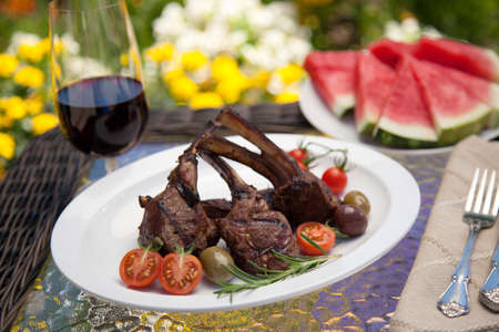 Grilled lamb chops served with cherry tomatoes and olives. Watermelon and wine. Outside dinner in a garden.の写真素材