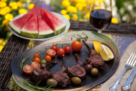 Grilled lamb chops served with cherry tomatoes, olives, and lemon. Watermelon and red wine. Outside dinner in a garden.の写真素材