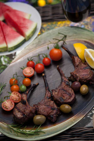 Grilled lamb chops served with cherry tomatoes, olives, and lemon. Watermelon and red wine. Outside dinner in a garden.の写真素材