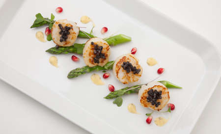Close up of delicious appetizer scallops with black caviar. Garnished with asparagus, pomegranate seeds, microgreens, and sauce. Elite food.の写真素材