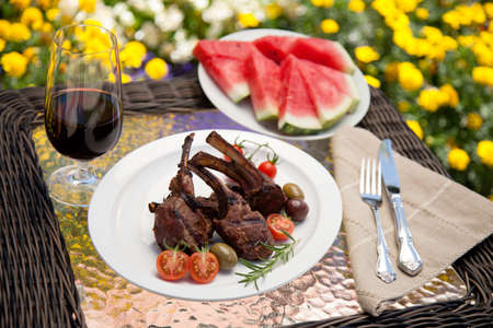 Grilled lamb chops served with cherry tomatoes and olives. Watermelon and wine. Outside dinner in a garden.の写真素材