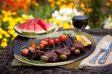 Grilled lamb chops served with cherry tomatoes and olives. Watermelon and wine. Outside dinner in a garden.の写真素材