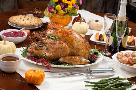 Roasted pepper turkey for Thanksgiving, garnished with pink pepper,  and fresh rosemary twigs on a dinner table decorated with mini pumpkins, beans, carrots, baked potato, pie, cranberry relish, gravy, flowers, candles, and flutes of champagne.の写真素材
