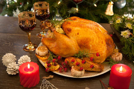 Rustic style roasted Christmas turkey garnished with roasted garlic, lemon, and rosehips. Surrounded with rustic Christmas ornaments, candles, wine, flowers, and Christmas tree in the background.の写真素材