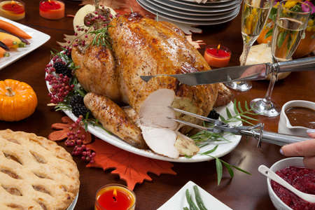 Carving roasted pepper turkey for Thanksgiving, garnished with pink pepper, black berry, and fresh rosemary twigs on a dinner table decorated with mini pumpkins, beans, carrots, baked potato, pie, cranberry relish, gravy, flowers, candles, and flutes of champagne.の写真素材