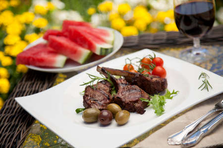 Grilled lamb chops served with cherry tomatoes and olives. Watermelon and wine. Outside dinner in a garden.の写真素材