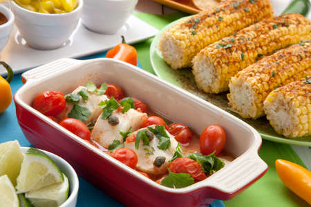 Mexican style baked cod with cherry tomatoes. Garnished with mexican condiments: guacamole, spicy salsa, marinated hot pepper, grilled corn, tortilla chips, and glass of beer.の写真素材