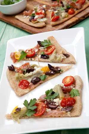 Closeup of a plate with few pieces of home made Mediterranean flatbread made with prosciutto, artichoke, cherry tomatoes, feta cheese, and olives. Garnished with parsley leafs.の写真素材