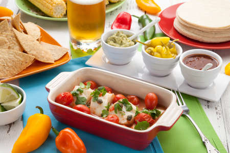 Mexican style baked cod with cherry tomatoes. Garnished with mexican condiments: guacamole, spicy salsa, marinated hot pepper, grilled corn, tortilla chips, and glass of beer.の写真素材