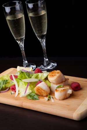 Close up of delicious savory fried scallops salad with fresh raspberry, basil, green lettuce, and red onion. Glass of Champagne. Over black background. Contemporary appetizers series.の写真素材