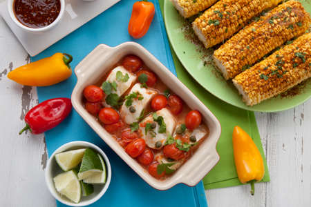 Mexican style baked cod with cherry tomatoes. Garnished with mexican condiments: guacamole, spicy salsa, marinated hot pepper, grilled corn, tortilla chips, and glass of beer.の写真素材