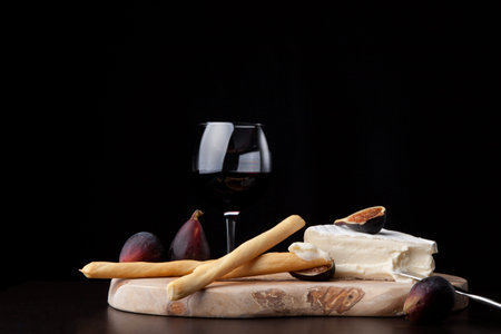 Closeup of Brie cheese with breadsticks, fresh black figs and glass of red wine - great snack for wine tasting. Over black background. Contemporary appetizers series.の写真素材