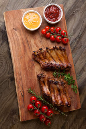 Grilled pork ribs with fresh cherry tomatoes, rosemary, oregano served with ketchup and mustard sauce on wooden board.の写真素材