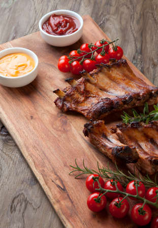 Grilled pork ribs with fresh cherry tomatoes, rosemary, oregano served with ketchup and mustard sauce on wooden board.の写真素材