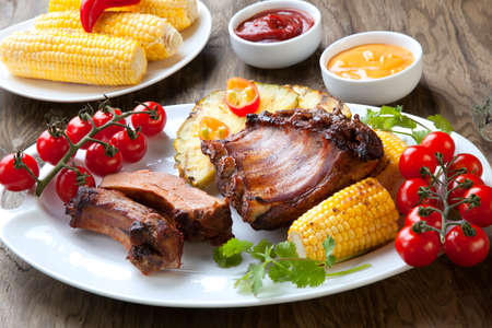 Grilled pork ribs, corn, and pineapple with chili pepper, cherry tomatoes, and sauces.の写真素材