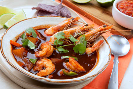 Closeup of a bowl of hot delicious spicy Mexican shrimp soup, garnished with whole shrimps and fresh cilantro. Limes, blue corn chips, salsa, guacamole, and marinated hot pepper.の写真素材