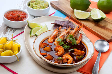 Closeup of a bowl of hot delicious spicy Mexican shrimp soup, garnished with whole shrimps and fresh cilantro. Limes, blue corn chips, salsa, guacamole, and marinated hot pepper.の写真素材