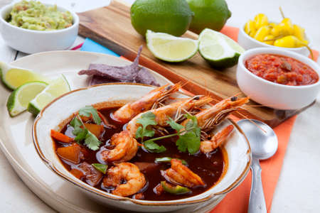 Closeup of a bowl of hot delicious spicy Mexican shrimp soup, garnished with whole shrimps and fresh cilantro. Limes, blue corn chips, salsa, guacamole, and marinated hot pepper.の写真素材