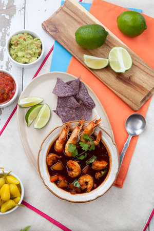 Closeup of a bowl of hot delicious spicy Mexican shrimp soup, garnished with whole shrimps and fresh cilantro. Limes, blue corn chips, salsa, guacamole, and marinated hot pepper.の写真素材
