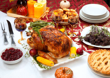 Garnished traditional fall roasted turkey garnished with corn on the cob, plums, and herbs. Served on a traditional decorated table with pumpkins, pie, green beans, baked vegetables, cranberry relish, corn bread, red wine, flowers, and candles.の写真素材