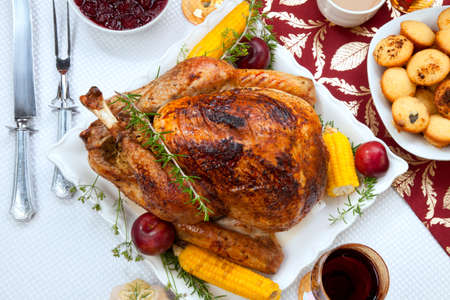 Garnished traditional fall roasted turkey garnished with corn on the cob, plums, and herbs. Served on a traditional decorated table with pumpkins, pie, green beans, baked vegetables, cranberry relish, corn bread, red wine, flowers, and candles.の写真素材