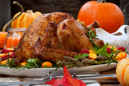 Citrus glazed roasted Turkey for Thanksgiving celebration garnished with kumquat, raspberry, asparagus, oregano, and fresh rosemary twigs. Red wine, side dishes, and gravy. Holiday table decorated with pumpkins, candels, gourds, and fall leaves.の写真素材
