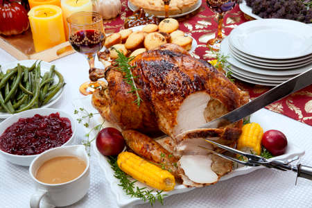 Carving garnished traditional fall roasted turkey garnished with corn on the cob, plums, and herbs. Served on a traditional decorated table with pumpkins, pie, green beans, baked vegetables, cranberry relish, corn bread, red wine, flowers, and candles.の写真素材