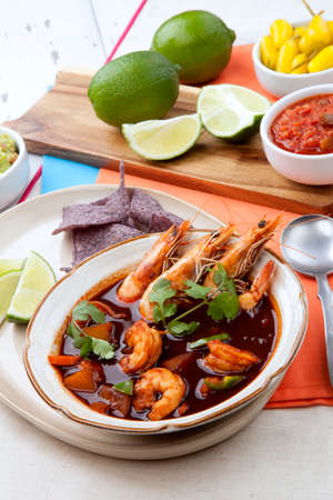 Closeup of a bowl of hot delicious spicy Mexican shrimp soup, garnished with whole shrimps and fresh cilantro. Limes, blue corn chips, salsa, guacamole, and marinated hot pepper.の写真素材