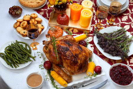 Garnished traditional fall roasted turkey garnished with corn on the cob, plums, and herbs. Served on a traditional decorated table with pumpkins, pie, green beans, baked vegetables, cranberry relish, corn bread, red wine, flowers, and candles.の写真素材