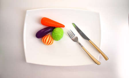 children's artificial vegetables, fork and knife, lie on a white plate on a white backgroundの写真素材