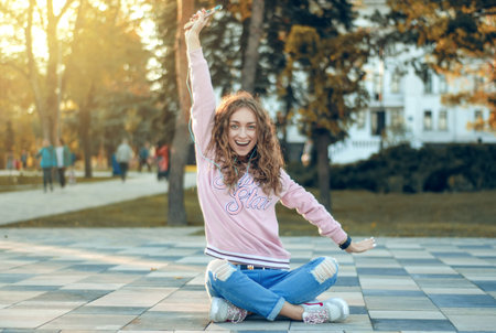 Woman sitting jeans sweatshirt sneakers watch smartphone mobile phone headphones selfie happy smiling on city streetの写真素材