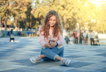Woman sitting jeans sweatshirt watch smartphone mobile phone headphones selfie on city street