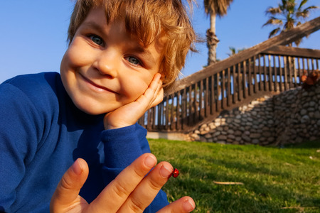 6 years old cute smiling boy, ladybug on his hand, sitting in the park, green grass and blue sky. Concept: connection between nature and people, family leisure, happy childhoodの写真素材