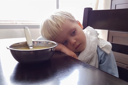 3-4 years old handsome blond boy with blue eyes sitting bored at the table near a plate of soup and does not want to eat. Concept: parenthood and childhoodの写真素材
