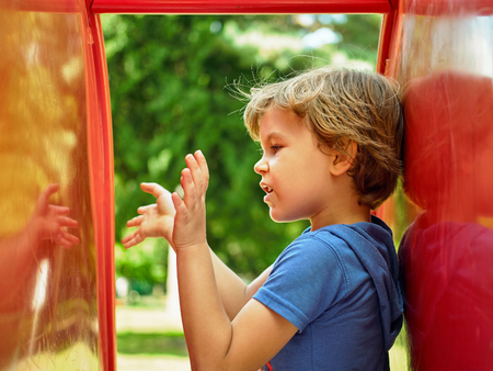 Cute little boy is playing with his reflection in childrens slide on the playground. Conducts a conversation with an imaginary friendの写真素材