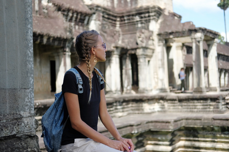 Young sun-tanned woman tourist with pigtails sitting near ancient temple. Man silhouette in background. Angkor Wat, Cambodiaの写真素材