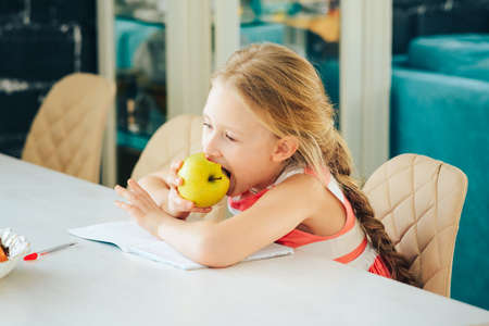 a little girl at the table with a notebook, doing homework and decided to eat a fresh Apple.の写真素材