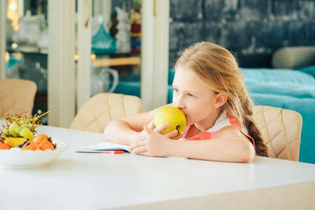 a little girl at the table with a notebook, doing homework and decided to eat a fresh Apple.の写真素材