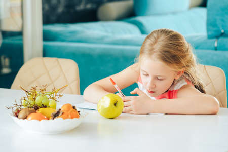 a little girl at the table with a notebook, doing homework and snacking on a fresh Apple.の写真素材