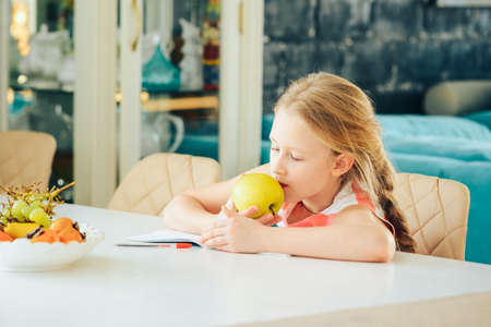 a little girl at the table with a notebook, doing homework and decided to eat a fresh Apple.の写真素材