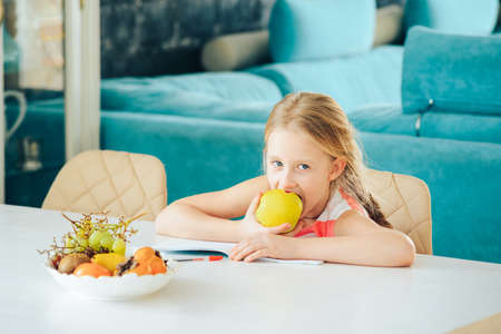 a little girl at the table with a notebook, doing homework and decided to eat a fresh Apple.の写真素材