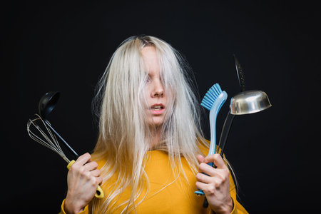A young girl with disheveled hair holds kitchen tools, arrives terrified of not being able to cook anything. The concept of a young housewife.の写真素材