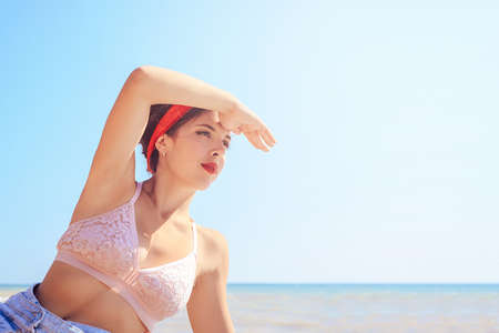 Closeup portrait, young sexy girl in Bra and denim skirt with a red bandage on his head. A young woman creates hand shadow before my eyes, and looking into the distance, on the coast of the ocean.の写真素材