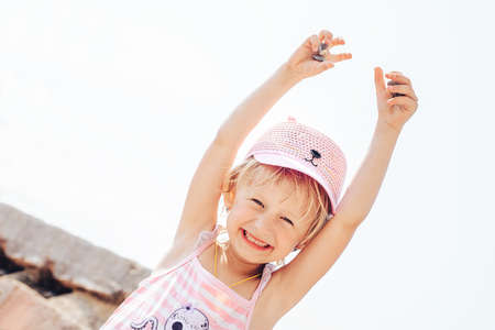 A cheerful smiling little girl in a rose hat raised her hands up playing with pebbles, on vacation by the seaの写真素材