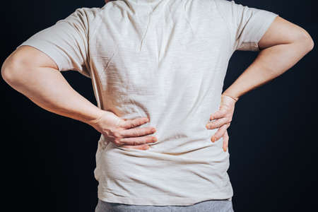 The guy put his hand on his sore back, standing against the black background. The concept of trauma, rheumatism, acute pain.の写真素材