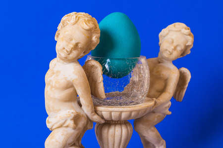 A statue of angels on a blue background holding a glass, cracked glass, inside an Easter egg turquoise. The concept of tradition of Easter.の写真素材