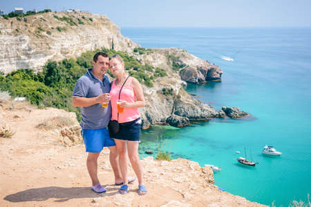 A young couple in love stands on top of a cliff overlooking a turquoise Bay with yachts, enjoying the views in their tourist adventure, sipping a cocktail from a clear glass.の写真素材