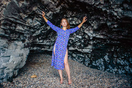 A young girl in a blue dress with polka dots hid in a cave, walking on the coast of the ocean. The concept of rest on the rocky coast.の写真素材